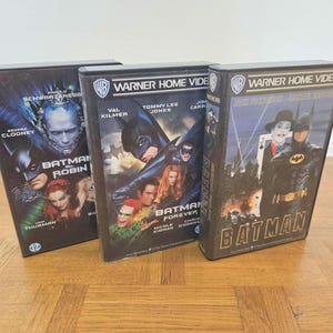 Può includere: Tre videocassette VHS vintage di Batman. Le videocassette presentano titoli di film come "Batman Forever" e "Batman Robin". Le videocassette hanno opere d'arte cinematografiche colorate e il logo Warner Home Video. Le videocassette sono esposte su una superficie di legno.