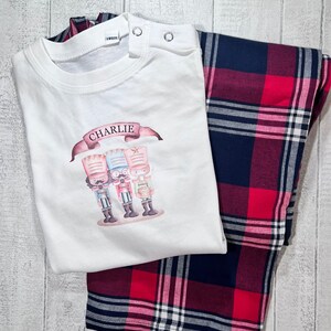 May include: White pyjamas with a red and blue plaid pattern. The top features a graphic of three nutcracker figures and the name "CHARLIE" in a banner.