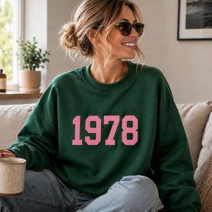 Puede incluir: Una sudadera de cuello redondo en color verde oscuro con el número 1978 impreso en una fuente grande, llamativa y de color rosa claro estilo universitario en el pecho. La prenda tiene mangas largas, cuello acanalado y un corte holgado.