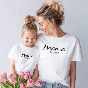 Puede incluir: Dos camisetas blancas con texto negro que dice "Mama EST. 2019" y "Mini EST. 2019". Una mujer y una niña pequeña llevan las camisetas. La niña sostiene un ramo de tulipanes rosas.