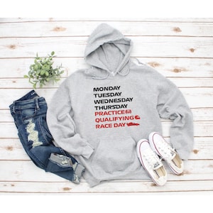Puede incluir: Una sudadera con capucha gris claro que presenta texto en negro MONDAY, TUESDAY, WEDNESDAY, THURSDAY y texto en rojo PRACTICE, QUALIFYING, RACE DAY, acompa&ntilde;ado de peque&ntilde;os iconos rojos de un coche, un casco y un coche de carreras.