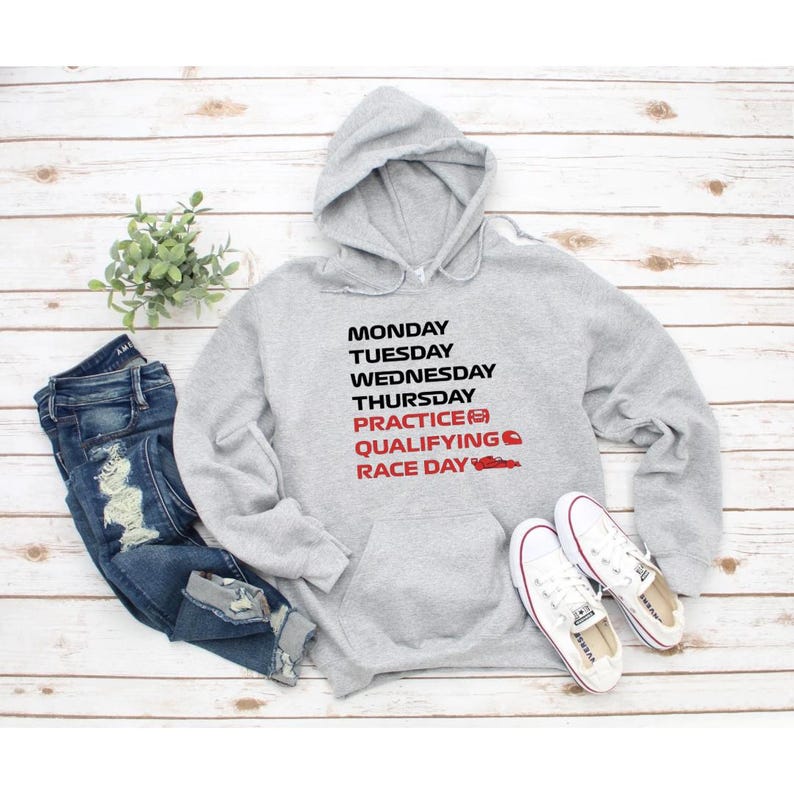 Puede incluir: Una sudadera con capucha gris claro que presenta texto en negro MONDAY, TUESDAY, WEDNESDAY, THURSDAY y texto en rojo PRACTICE, QUALIFYING, RACE DAY, acompa&ntilde;ado de peque&ntilde;os iconos rojos de un coche, un casco y un coche de carreras.