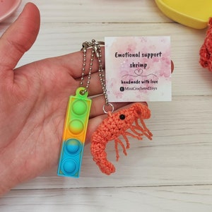 May include: A keychain with a crocheted orange shrimp and a multi-colored pop-it toy. The keychain is attached to a silver chain. The card attached to the keychain reads "Emotional support shrimp" and "handmade with love" with the Instagram handle "MiniCrochetedToys".