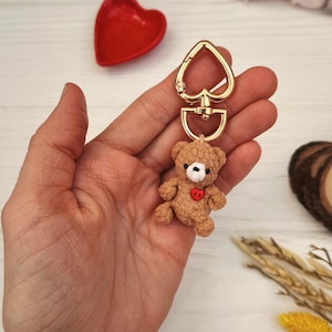 Może przedstawiać: Brązowy brelok do kluczy z misiem, ze złotym zapięciem w kształcie serca. Miś ma czerwone serce na piersi. W tle czerwone naczynie w kształcie serca.