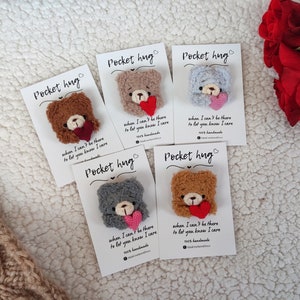 May include: Five small plush teddy bear pocket hug ornaments on white cards with the text "Pocket hug" and "when I can't be there to let you know I care". The bears are brown, grey, and light brown and are holding a red heart.