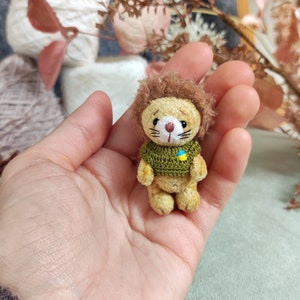 Könnte beinhalten: Ein kleiner gehäkelter Löwe, der einen grünen Pullover mit einem gelben und blauen Patch trägt. Der Löwe wird in der Hand einer Person gehalten.