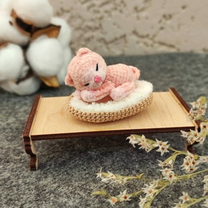 May include: A pink crocheted cat toy is sleeping on a small wooden bed with a white crocheted pillow. The bed is on a gray surface with white flowers.
