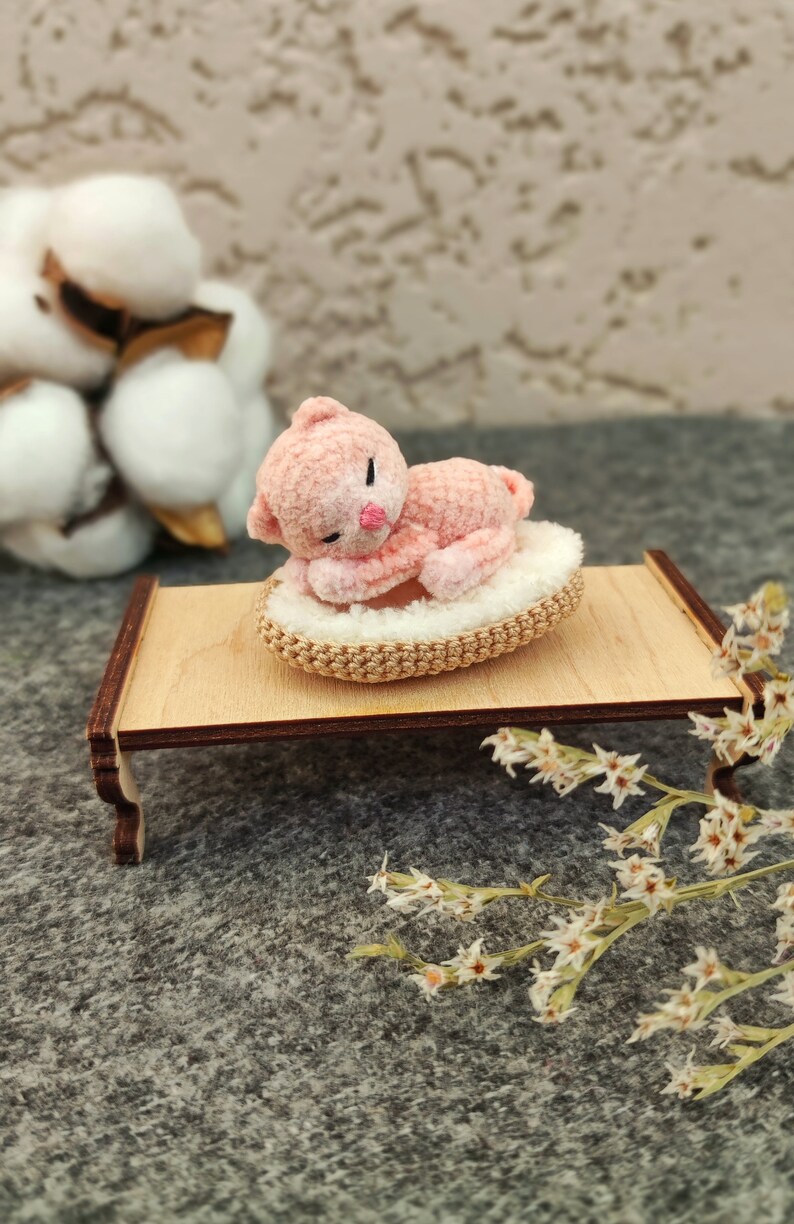 May include: A pink crocheted cat toy is sleeping on a small wooden bed with a white crocheted pillow. The bed is on a gray surface with white flowers.