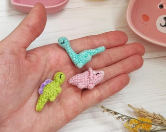 May include: Three small crocheted dinosaur toys. The dinosaurs are green, blue, and pink. They are sitting on a person's hand.