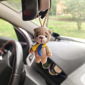 May include: A small brown teddy bear with a yellow and blue scarf hanging from the rearview mirror of a car.