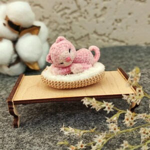 May include: A pink crocheted cat toy laying on a crocheted bed, on a small wooden table. The table is on a gray surface with white flowers.