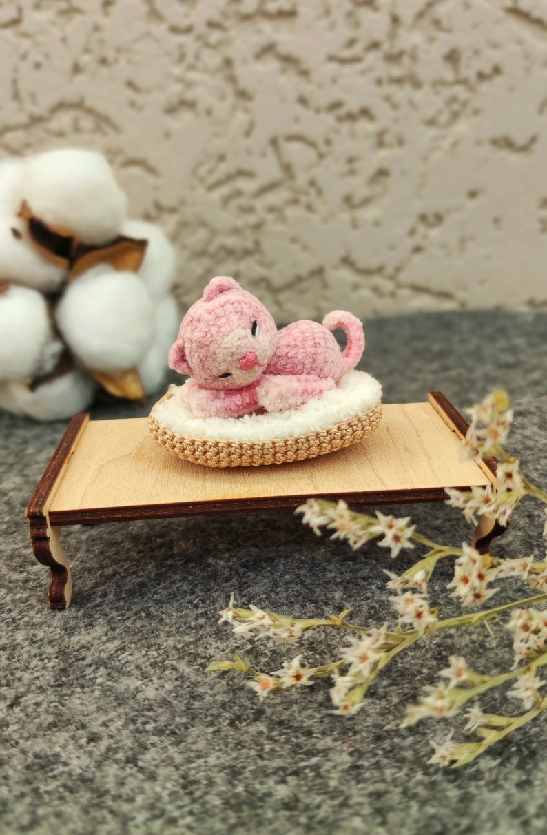 May include: A pink crocheted cat toy laying on a crocheted bed, on a small wooden table. The table is on a gray surface with white flowers.