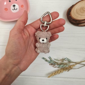 May include: A light brown teddy bear keyring. The bear is made of a soft, plush material and has a white muzzle and black eyes. It is attached to a silver-coloured heart-shaped clasp.