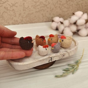 May include: A selection of small, crocheted chicken figurines in shades of brown, beige, white, and grey, each with a red comb. The chickens are displayed on a decorative tray. One chicken is held in a hand.