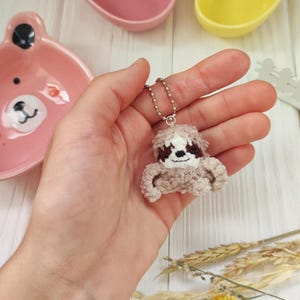 Puede incluir: Un pequeño llavero de perezoso de crochet marrón con una cadena plateada. El perezoso está colgado de una mano.