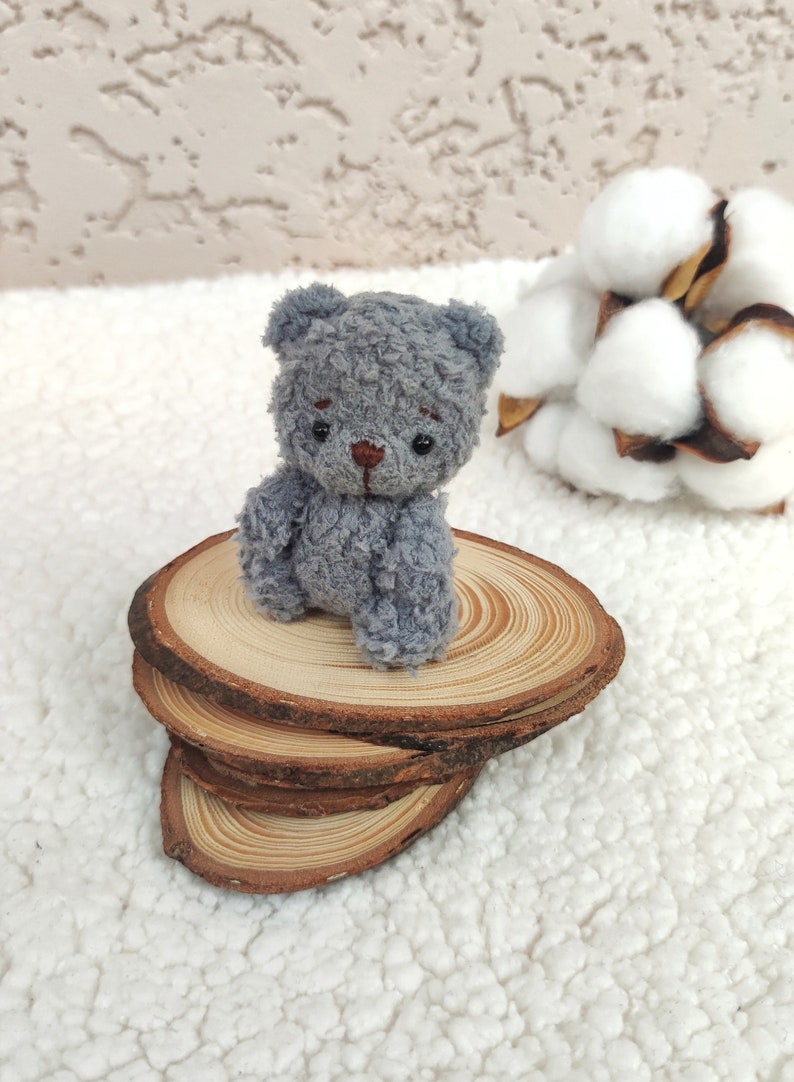 K&ouml;nnte beinhalten: Ein kleiner grauer Teddyb&auml;r sitzt auf einem Stapel aus Holzscheiben. Der B&auml;r hat eine braune Nase und schwarze Augen. Die Holzscheiben sind in einem Stapel von drei angeordnet.