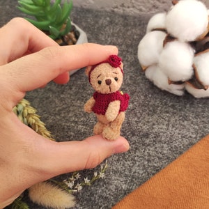 May include: A small, brown teddy bear wearing a red crocheted dress and a red flower on its head. The bear is being held by a hand.