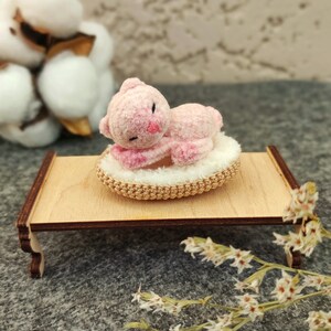 May include: A pink crocheted cat toy is sleeping on a small, round, crocheted bed. The bed is on a wooden stand with a light brown finish. The stand has two legs on each side.