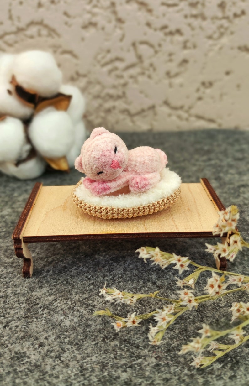 May include: A pink crocheted cat toy is sleeping on a small, round, crocheted bed. The bed is on a wooden stand with a light brown finish. The stand has two legs on each side.
