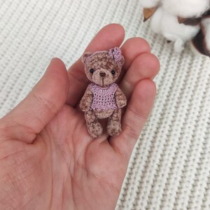 May include: A small brown crocheted teddy bear wearing a pink crocheted dress and a pink flower headband.