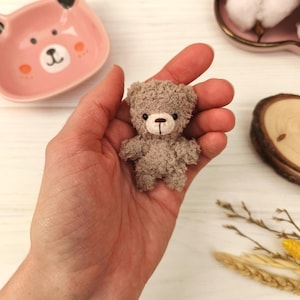 May include: A small, handmade teddy bear in a person's hand. The bear is light brown with a textured appearance, black eyes, and a brown nose. A pink bear-shaped dish is in the background, along with decorative elements.