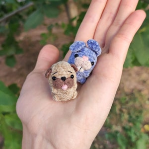 May include: A hand holding two small crocheted stuffed animals. One is a blue bunny with long ears and the other is a brown pug with a pink tongue sticking out.
