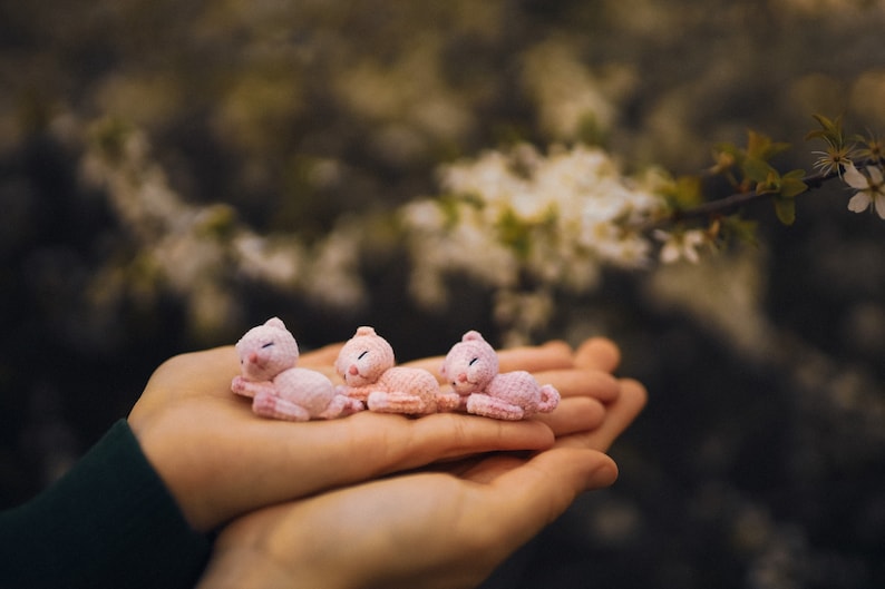 May include: Four small pink crocheted animal figurines, possibly kittens, are resting in the palm of a person's hand. The figurines are arranged in a row, with their heads facing the viewer. The background is blurred and features a tree branch with white blossoms.