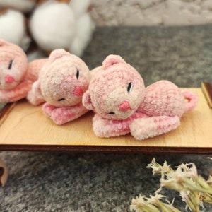 May include: Three pink plush toy kittens lying on a wooden surface. The kittens have embroidered eyes and noses.