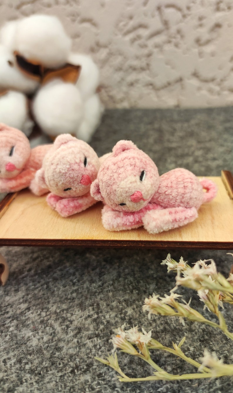 May include: Three pink plush toy kittens lying on a wooden surface. The kittens have embroidered eyes and noses.