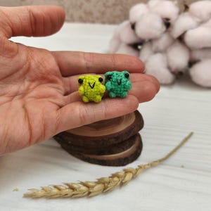May include: Two tiny, handmade crochet frogs. One is lime green, the other is teal. Both have black eyes and a small, stitched smile. The frogs are displayed on a wooden slice, with a wheat stalk and cotton buds in the background.
