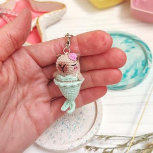May include: A crocheted mermaid keychain with a pink flower on its head. The mermaid is light brown with a teal tail and is holding a small pink flower.
