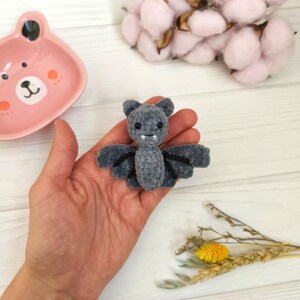 May include: A gray crocheted bat with outstretched wings. The bat has small ears, a smiling face, and black accents. The bat is held in a hand, with a pink bear-shaped dish and cotton buds in the background.