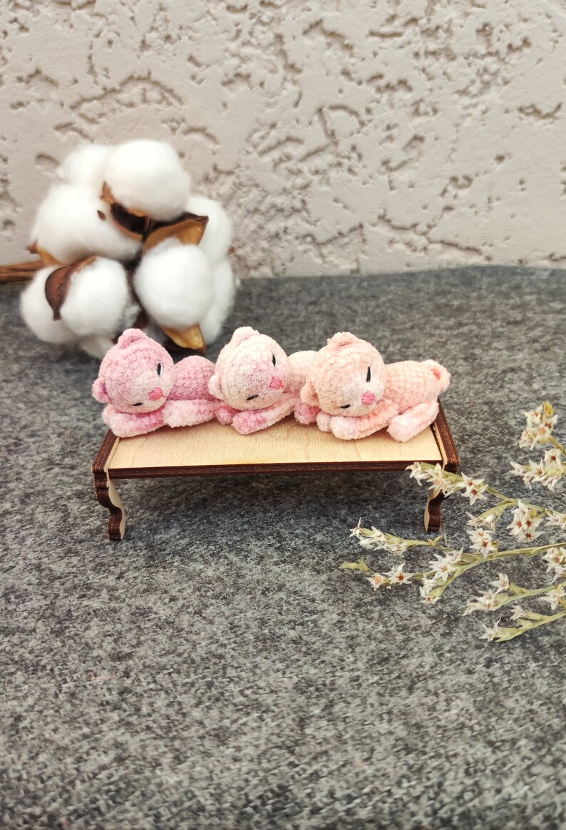 May include: Four pink crocheted kittens laying on a small wooden table. The kittens are facing the same direction and have black eyes and noses.