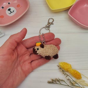 Peut inclure: Un porte-clés en crochet marron et blanc en forme d'animal avec un ananas jaune et vert sur le dessus. Le porte-clés est attaché à un anneau de clé argenté.