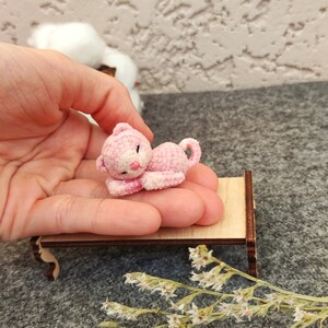 May include: A small pink crocheted cat laying on a miniature wooden bed. The cat has a white face and pink ears. The bed is on a gray surface with dried white flowers.