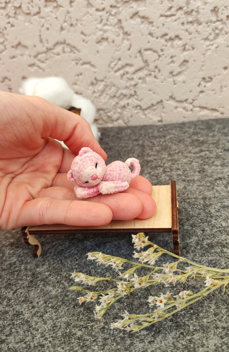May include: A small pink crocheted cat laying on a miniature wooden bed. The cat has a white face and pink ears. The bed is on a gray surface with dried white flowers.