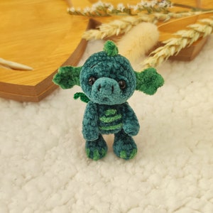 May include: A small, green crocheted dragon with a light green stripe down its back. The dragon has large, black eyes and a small, green tail.