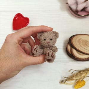 Puede incluir: Un pequeño oso de peluche hecho a mano, en tonos marrones, sostenido en una mano. El oso tiene detalles marrón oscuro en sus patas y una nariz cosida. La imagen también incluye un plato rojo en forma de corazón, algodón y rodajas de madera.