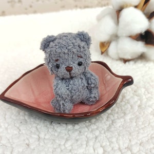 K&ouml;nnte beinhalten: Ein kleiner grauer Teddyb&auml;r mit braunen Augen sitzt in einer rosa, blattartigen Schale. Die Schale steht auf einer wei&szlig;en, flauschigen Oberfl&auml;che.