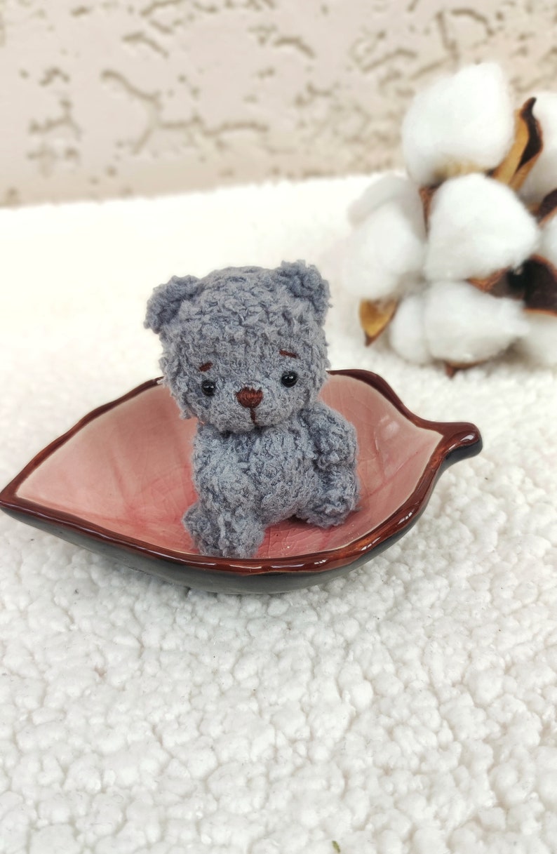 K&ouml;nnte beinhalten: Ein kleiner grauer Teddyb&auml;r mit braunen Augen sitzt in einer rosa, blattartigen Schale. Die Schale steht auf einer wei&szlig;en, flauschigen Oberfl&auml;che.