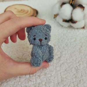 K&ouml;nnte beinhalten: Ein kleiner, grauer, Pl&uuml;schteddyb&auml;r mit schwarzen Augen und einer braunen Nase. Der B&auml;r sitzt auf einer wei&szlig;en, flauschigen Oberfl&auml;che.