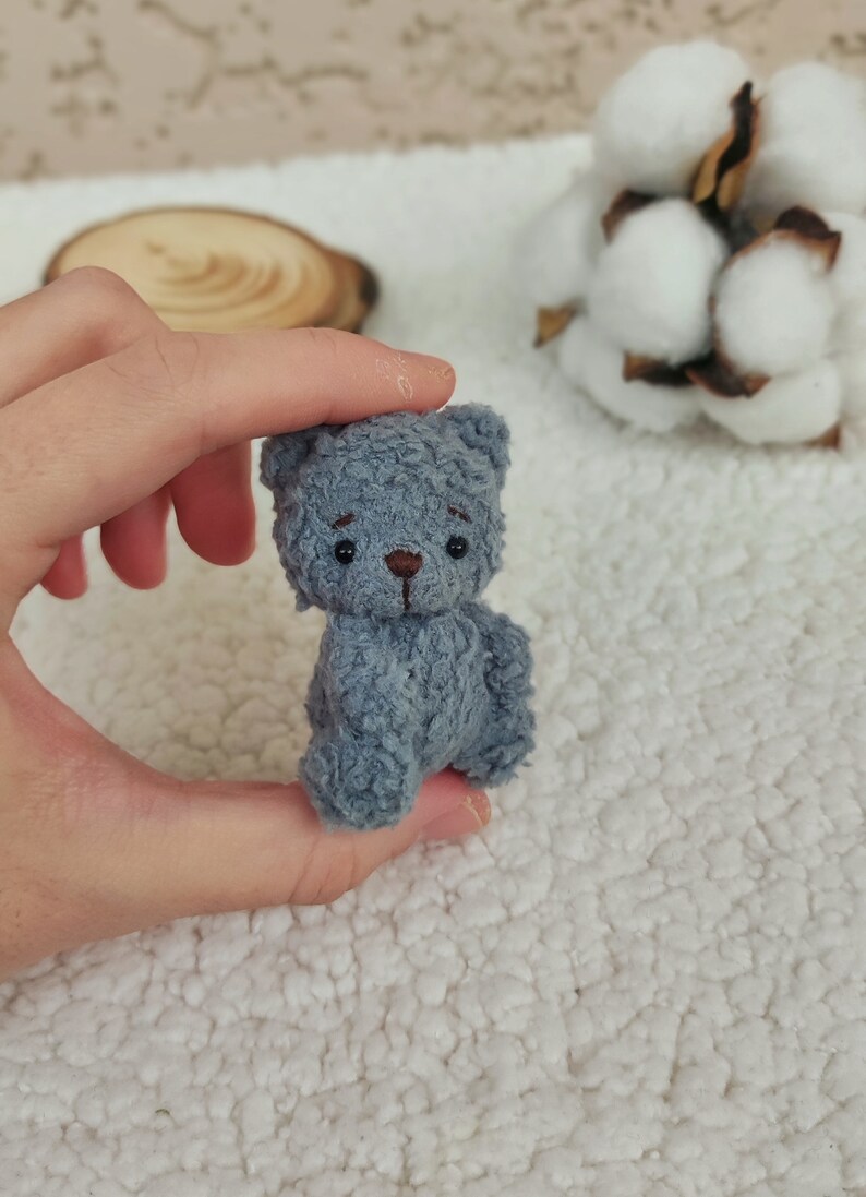K&ouml;nnte beinhalten: Ein kleiner, grauer, Pl&uuml;schteddyb&auml;r mit schwarzen Augen und einer braunen Nase. Der B&auml;r sitzt auf einer wei&szlig;en, flauschigen Oberfl&auml;che.