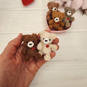 Może przedstawiać: Ręka trzyma dwa małe, ręcznie robione misie, jeden brązowy i jeden kremowy, każdy z czarnym guzikiem i czerwonym sercem. Widoczna jest również mała czerwona miseczka w kształcie serca oraz grupa misiów w różowej miseczce w kształcie serca.