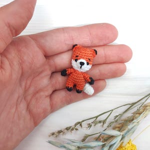 Puede incluir: Un pequeño juguete de zorro de crochet con pelaje naranja, la punta de la cola blanca y ojos negros. El zorro está sentado en una mano.