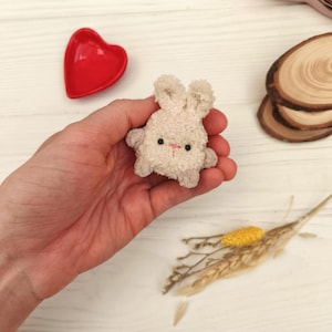 Peut inclure: Un petit jouet en forme de lapin en peluche de couleur beige clair, avec des yeux noirs et un nez rose. Le lapin est tenu dans une main, avec un plat en forme de cœur rouge et des tranches de bois décoratives en arrière-plan. C'est un article mignon et fait à la main.