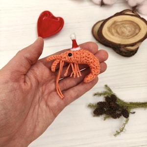 Puede incluir: Un camarón naranja tejido a ganchillo con un pequeño gorro de Papá Noel. El camarón tiene ojos negros y largas antenas naranjas. El camarón se sostiene en una mano, con un plato rojo en forma de corazón, rodajas de madera y vegetación en el fondo.