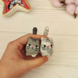Könnte beinhalten: Zwei kleine gehäkelte graue und weiße Katzenspielzeuge mit schwarzen Augen und rosa Nasen. Die Katzenspielzeuge werden in der Hand einer Person gehalten.