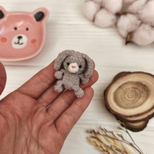 May include: A small, gray crocheted bunny with floppy ears and a pink nose. The handmade toy is held in a hand, with a pink bear-shaped dish and cotton buds in the background. The bunny is a charming, miniature creation.