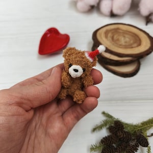 Könnte beinhalten: Ein kleiner brauner Teddybär mit rotem und weißem Weihnachtsmannhut. Der Bär hat schwarze Augen und eine weiße Schnauze. Der Bär wird in einer Hand gehalten, mit einer roten herzförmigen Schale und Holzscheiben im Hintergrund.