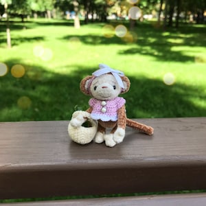 May include: A small crocheted monkey doll wearing a pink and white dress and a white bow. The monkey is holding a white crocheted basket and is sitting on a wooden bench.